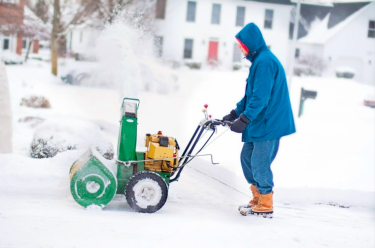Persona abrigada con ropa de invierno utilizando un quitanieves para despejar un camino cubierto de nieve en una zona residencial